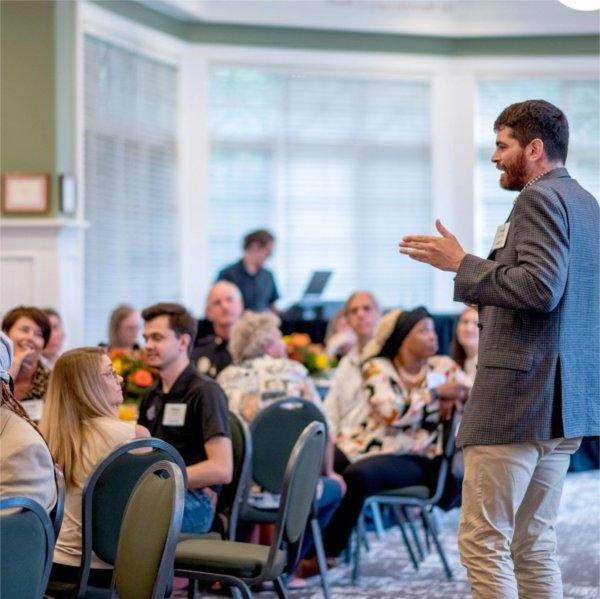 man stands and addresses room of people seated at round tables in Alumni House